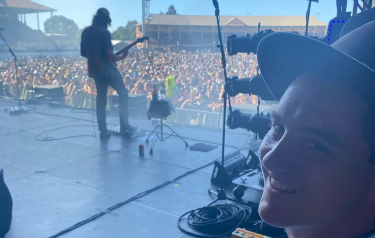 ABC reporter Charles Brice smiling backstage at a music festival, with a guitarist performing on stage in front of a large outdoor crowd behind him.